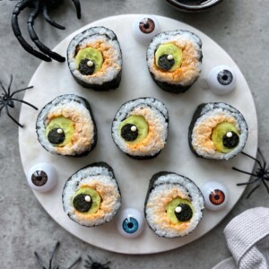 A round white plate holds six sushi rolls decorated to look like eyeballs, surrounded by plastic eyeballs and black plastic spiders on a gray surface, making these festive Halloween bites a standout among spooky food recipes.