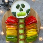 A Halloween-themed veggie platter arranged as a skeleton makes for one of the healthiest bites—bell pepper ribs, cucumber spine, pretzel joints, and a dip bowl skull with cucumber features. A plastic spider adds spooky fun to this festive food recipe.
