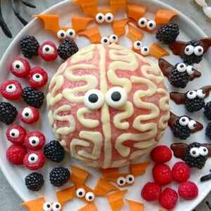 A plate with a brain-shaped dessert topped with candy eyes and icing, surrounded by berries and carrot pieces decorated with googly eyes, makes for playful monster-inspired bites perfect for fun food recipes.