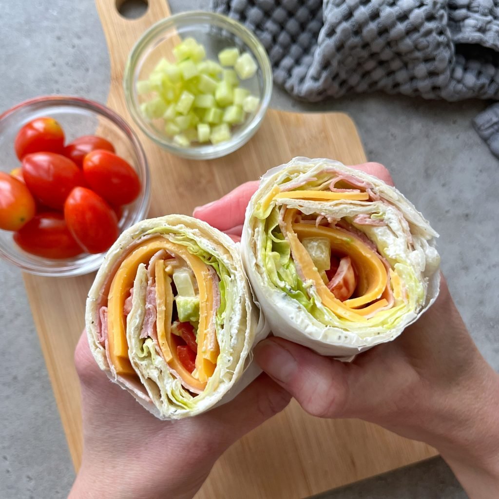 Two hands hold a sliced Ham and Cheese Salad Wrap filled with ham, cheese, lettuce, and tomato. In the background, a wooden board holds a bowl of grape tomatoes and chopped cucumber. A gray cloth is partially visible.