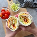 Two hands hold a Ham and Cheese Salad Wrap filled with ham, cheese, lettuce, and tomato. In the background, a wooden board displays grape tomatoes and a bowl of diced cucumber atop a gray textured cloth.