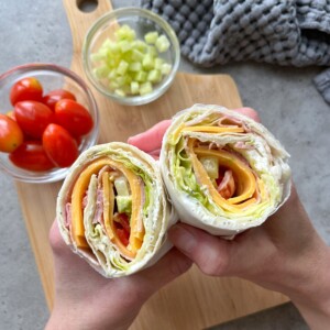 Two hands hold a Ham and Cheese Salad Wrap filled with ham, cheese, lettuce, and tomato. In the background, a wooden board displays grape tomatoes and a bowl of diced cucumber atop a gray textured cloth.