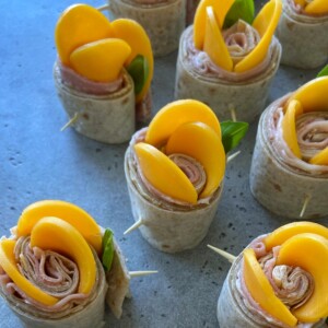 Small wrap rolls filled with ham are arranged upright, garnished with yellow peach slices and fresh green basil leaves for healthy bites—each secured with a toothpick, on a gray countertop—perfect for your food recipes collection.