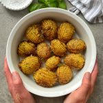 A pair of hands holds a white bowl filled with twelve hasselback baked potatoes, garnished with herbs. The bowl rests on a gray surface with a striped cloth, fresh basil leaves, and a dish of salt nearby.