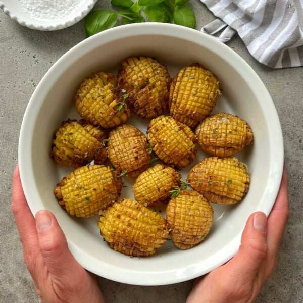 A pair of hands holds a white bowl filled with twelve hasselback baked potatoes, garnished with herbs. The bowl rests on a gray surface with a striped cloth, fresh basil leaves, and a dish of salt nearby.