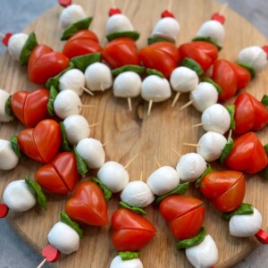 Valentine-themed skewers arranged in a heart shape on a wooden board, featuring cherry tomatoes cut into hearts, fresh basil leaves, and mozzarella balls.