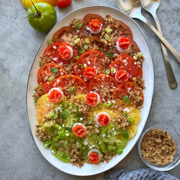 An oval platter with sliced red and yellow heirloom tomatoes, topped with chopped green onions, small dollops of a red spread, and sprinkled with breadcrumbs. Spoons and extra breadcrumbs are nearby on a gray surface.