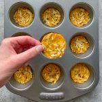 A hand holds a round, baked homemade cheese cracker above a muffin tin filled with similar cheese crisps, each topped with shredded cheese and herbs.