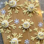 Round cookies decorated as cute snowflakes with wide eyes and yellow arms, arranged on a wooden board alongside white paper snowflakes and small string lights—perfect little bites for winter gatherings or festive food recipes.