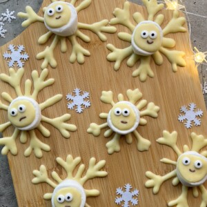 Round cookies decorated as cute snowflakes with wide eyes and yellow arms, arranged on a wooden board alongside white paper snowflakes and small string lights—perfect little bites for winter gatherings or festive food recipes.