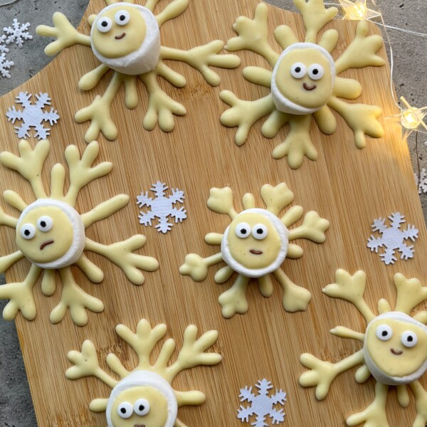 Round cookies decorated as cute snowflakes with wide eyes and yellow arms, arranged on a wooden board alongside white paper snowflakes and small string lights—perfect little bites for winter gatherings or festive food recipes.