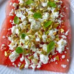 A slice of hot honey watermelon carpaccio is topped with crumbled feta, chopped pistachios, and fresh green basil leaves, served on a white plate.