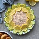A round plate of creamy Hot Smoked Salmon Dip is garnished with alternating slices of lemon and cucumber in a circular pattern. A blue napkin, lemon halves, and toasted bread slices are nearby on a gray surface.