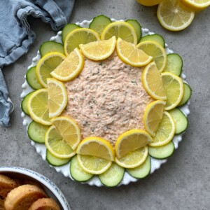 A round plate of creamy Hot Smoked Salmon Dip is garnished with alternating slices of lemon and cucumber in a circular pattern. A blue napkin, lemon halves, and toasted bread slices are nearby on a gray surface.