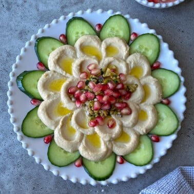 A beautifully arranged hummus platter takes center stage, artfully shaped like a flower and topped with olive oil, chopped pickles, and pomegranate seeds, all surrounded by crisp cucumber slices on a white decorative plate.