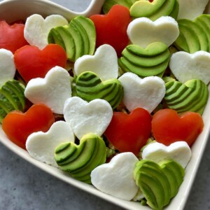 A heart-shaped bowl filled with heart-shaped slices of tomato, mozzarella cheese, and avocado—perfect for health-conscious bites—arranged on a gray surface.