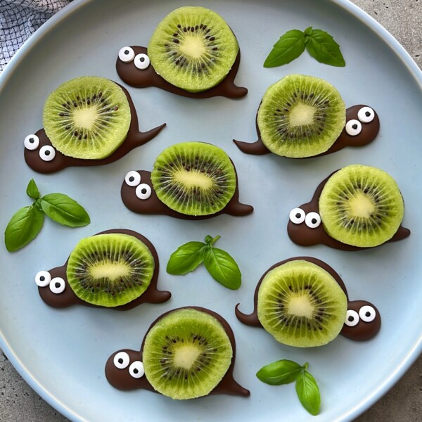 Sliced kiwis arranged on a plate, each with a chocolate base shaped like a snail’s head and tail, candy eyes, and fresh basil leaves for decoration—these playful snail-themed bites are perfect for fun food recipes.