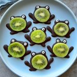 Seven frog-shaped bites made with chocolate for the bodies and legs, white and black candy eyes, and slices of kiwi for faces, arranged on a light blue plate—perfect for fun food recipes or healthy party dishes.