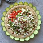 A circular spread of layered hummus dip topped with chopped tomatoes, cucumbers, feta cheese, herbs, and nuts, surrounded by overlapping cucumber slices on a gray surface.