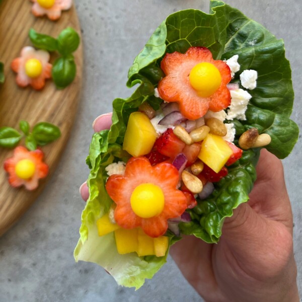 A hand holds a lettuce leaf filled with colorful, health-focused bites—flower-shaped strawberries and carrots, yellow mango, feta cheese, pine nuts, and red onion. A wooden board with more flower-shaped toppings sits in the background.