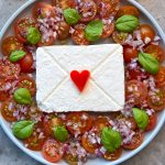 A plate with cherry tomato slices, chopped red onions, and basil leaves arranged around a block of white cheese decorated like an envelope, topped with a red heart-shaped piece of vegetable—perfect for health-conscious bites.