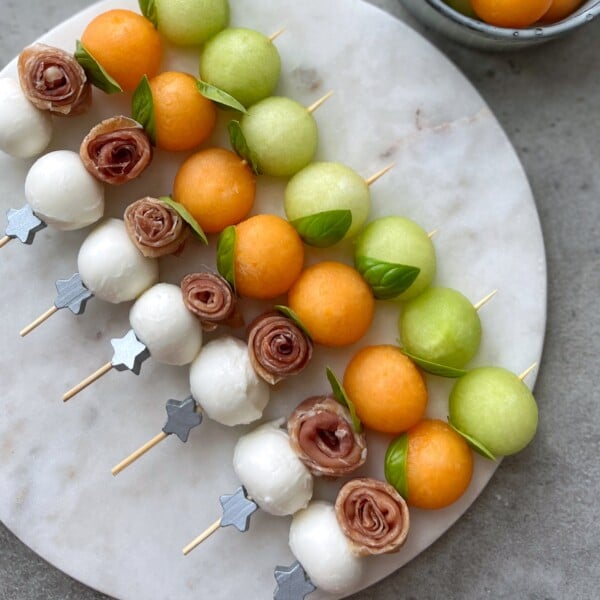 Skewers with mozzarella balls, rolled prosciutto, green and orange melon balls, and fresh basil leaves, arranged neatly on a round marble platter—perfect for healthy bites or inspiring new food recipes.