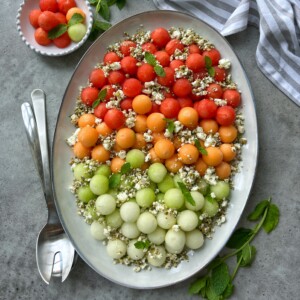 An oval platter of melon feta salad features rows of red, orange, and green melon balls topped with crumbled cheese, herbs, and seeds. Fresh mint leaves, serving utensils, a striped napkin, and a bowl of extra melon balls complete the scene.