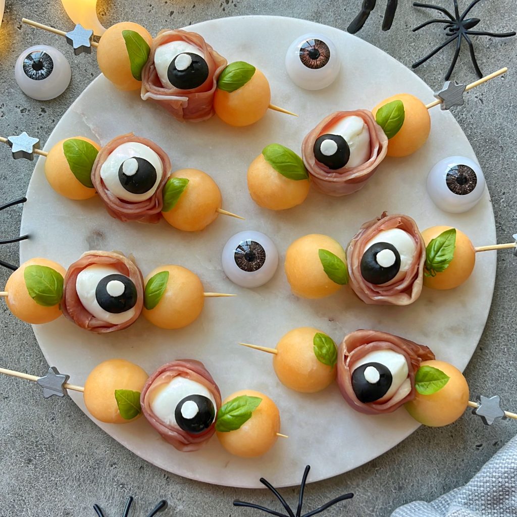 A round platter with skewers of melon balls, mozzarella, prosciutto, and basil arranged to resemble eyeballs—these spooky bites are perfect for Halloween food recipes, surrounded by fake eyeballs and plastic spiders.