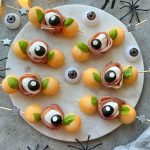 A round platter with skewers of melon balls, mozzarella, prosciutto, and basil arranged to resemble eyeballs—these spooky bites are perfect for Halloween food recipes, surrounded by fake eyeballs and plastic spiders.