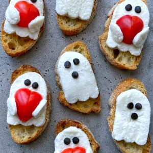 Slices of bread topped with mozzarella cheese ghosts—some holding red tomato hearts—all have olive piece eyes. These fun bites make a spooky addition to your food recipes and health-conscious dishes, arranged on a grey surface.