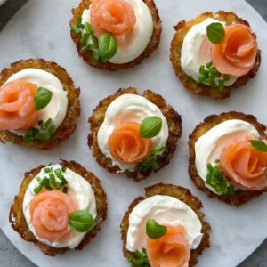 Seven potato latkes topped with dollops of sour cream, slices of smoked salmon shaped like roses, chopped chives, and fresh basil leaves. Arranged elegantly on a round white plate.