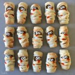 Rows of baked mummy bites, wrapped in dough with chocolate centers, candy eyes, and red ketchup mouths, arranged on a gray surface—perfect for fun food recipes or spooky party dishes.