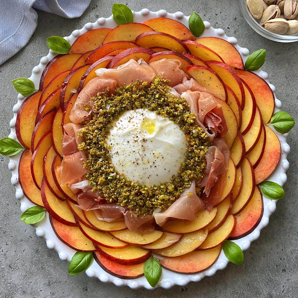 A round white plate with concentric layers of sliced peaches and nectarines, prosciutto, and crushed pistachios surrounding a ball of burrata cheese, garnished with basil leaves and a drizzle of olive oil—a refreshing nectarine salad.