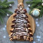 A Nutella Puff Pastry Christmas Tree decorated with chocolate drizzle and powdered sugar, displayed on a wooden board, surrounded by fir branches, ornaments, and snowflake decorations.
