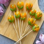 Eight olive appetizer sticks with yellow cherry tomatoes and green leaves, arranged to look like tulips, are placed on a wooden board, with pink flowers and a pink ribbon nearby.