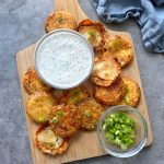 A wooden board with parmesan chips with onion, crispy and golden, topped with sliced green onions, a bowl of creamy white dip with herbs, and extra green onions on the side, arranged on a gray surface with a blue cloth nearby.
