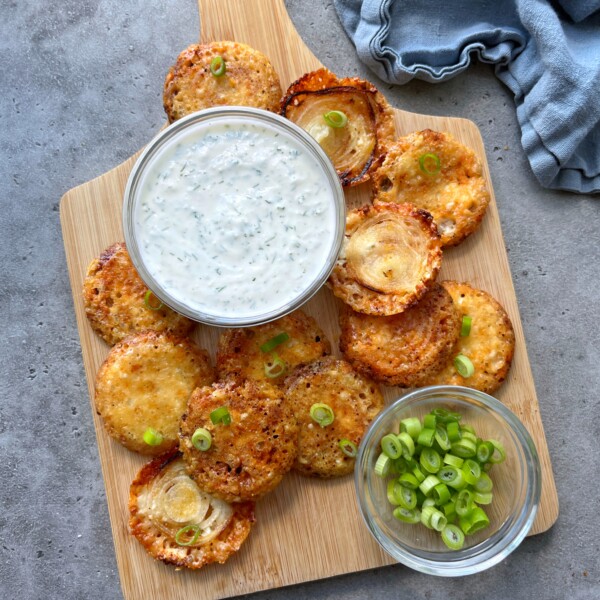 A wooden board with parmesan chips with onion, crispy and golden, topped with sliced green onions, a bowl of creamy white dip with herbs, and extra green onions on the side, arranged on a gray surface with a blue cloth nearby.