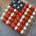 Skewers with cherry tomatoes, mozzarella balls, blueberries, and small cheese stars are arranged on a wooden board to resemble the American flag—perfect for healthy bites at festive gatherings or as fun additions to your food recipes.
