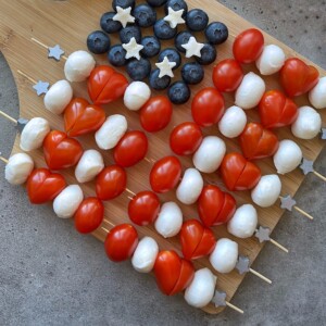 Skewers with cherry tomatoes, mozzarella balls, blueberries, and small cheese stars are arranged on a wooden board to resemble the American flag—perfect for healthy bites at festive gatherings or as fun additions to your food recipes.