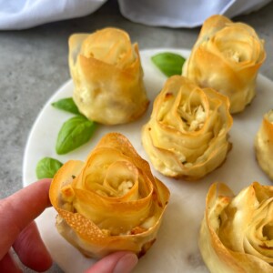 A hand holds a baked pastry shaped like a rose on a white plate, surrounded by more phyllo appetizers and fresh basil leaves. The pastries have golden, crisp edges and a creamy filling.