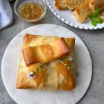 A golden-brown phyllo baked brie, wrapped like a present and garnished with fresh herbs, sits on a marble plate. Nearby are a bowl of jelly and slices of toasted bread on a white plate.
