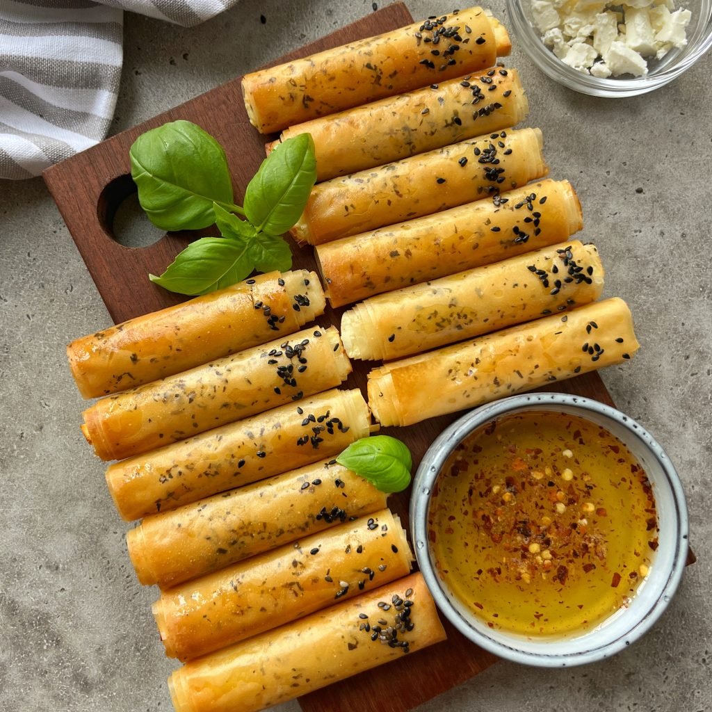 Golden, crispy Phyllo Feta Fries rolled and topped with black sesame seeds are arranged on a wooden board with basil leaves. A small bowl of seasoned olive oil and a bowl of crumbled cheese are nearby.