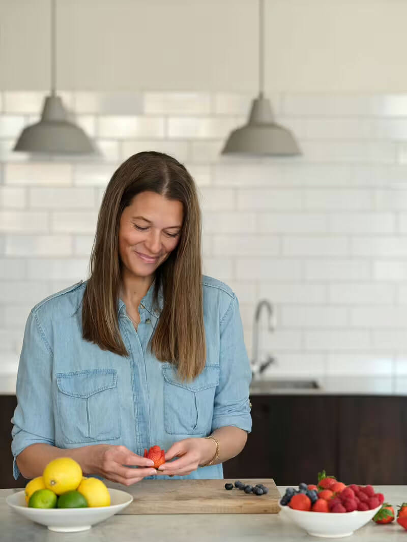 A woman with long brown hair, wearing a light denim shirt, smiles while preparing fruit at a kitchen counter. There are lemons on a plate and berries on a cutting board in front of her. White tiles and hanging lights are in the background.