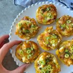 A hand reaches for a plate of six baked, cheese-topped potato bites garnished with chopped green onions and herbs, displayed on a white, scalloped-edge plate—a delightful addition to your favorite food recipes.