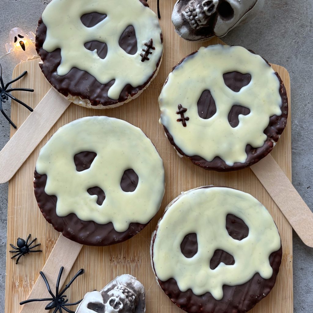 Four Poison Apple Rice Cakes, covered in chocolate and decorated with white chocolate skull faces on sticks, are arranged on a wooden board surrounded by toy spiders, skull decorations, and fairy lights for a spooky Halloween vibe.
