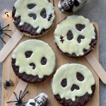 Four Poison Apple Rice Cakes, covered in chocolate and decorated with white chocolate skull faces on sticks, are arranged on a wooden board surrounded by toy spiders, skull decorations, and fairy lights for a spooky Halloween vibe.