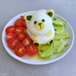 A plate with a cute bear shape made from white cheese, decorated with cucumber and cheese pieces, surrounded by sliced cherry tomatoes and fish-shaped cucumber bites—an adorable dish perfect for healthy food recipes, all drizzled with olive oil.