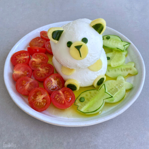 A plate featuring a bear-shaped sculpture made of white cheese, with cucumber and cheese details, surrounded by sliced cherry tomatoes and cucumber fish shapes—fun food bites perfect for health-focused dishes, all drizzled with olive oil.