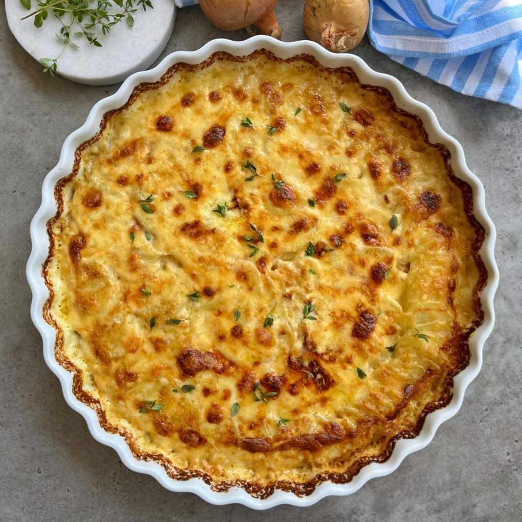 A golden-brown potato gratin, a classic among comforting food recipes, baked in a white scalloped dish and garnished with fresh herbs, sits on a grey countertop near mushrooms, a blue-striped towel, and a sprig of thyme.