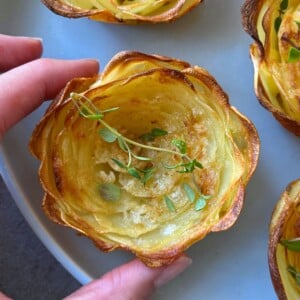 A hand holds a crispy, golden-brown potato rose, garnished with fresh thyme and displayed on a light blue plate with more elegant potato roses in the background.